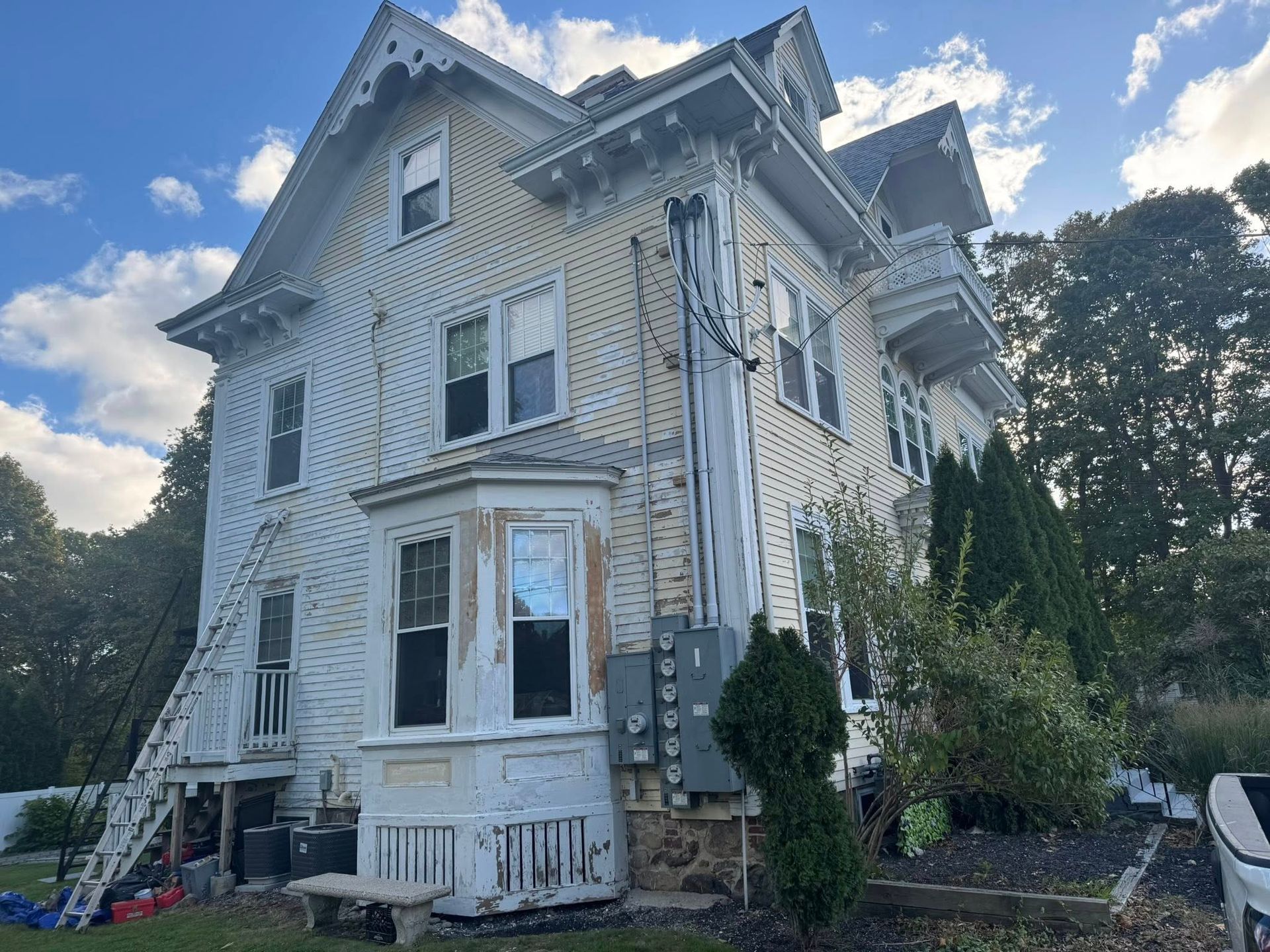 Weathered white Victorian house with bay windows and a front porch, viewed from the corner under a cloudy sky