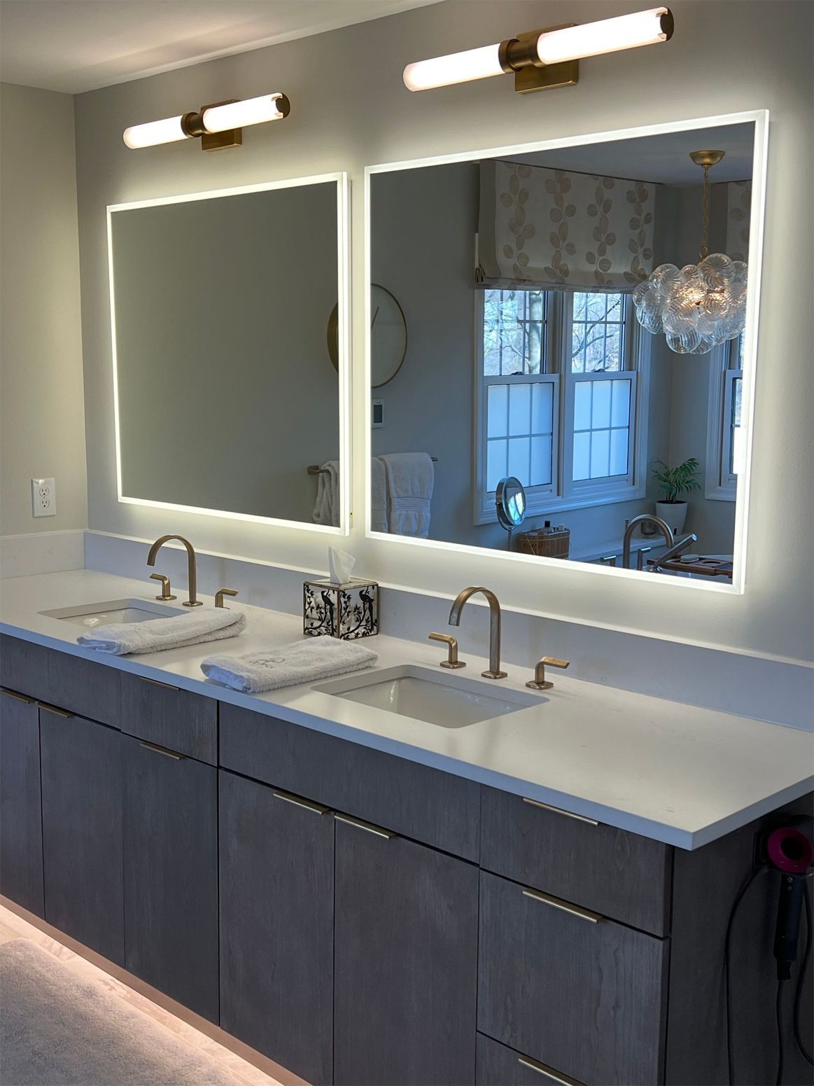 Bathroom with two rectangular mirrors with built-in lights over a double vanity, gold fixtures, and gray cabinets.