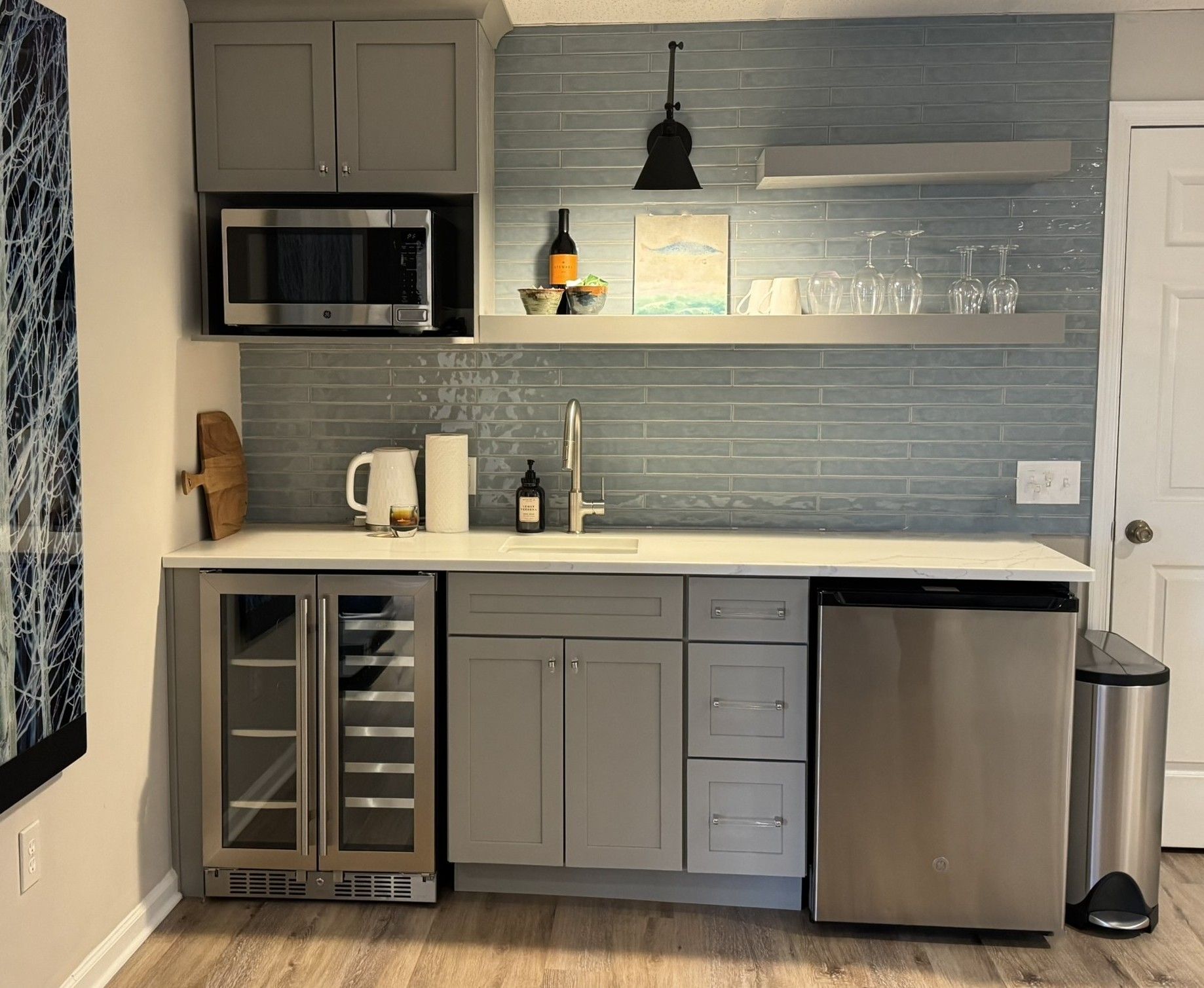 Compact gray kitchenette with wine fridge, microwave, sink, and dishwasher on wood floor, against blue tiled backsplash.