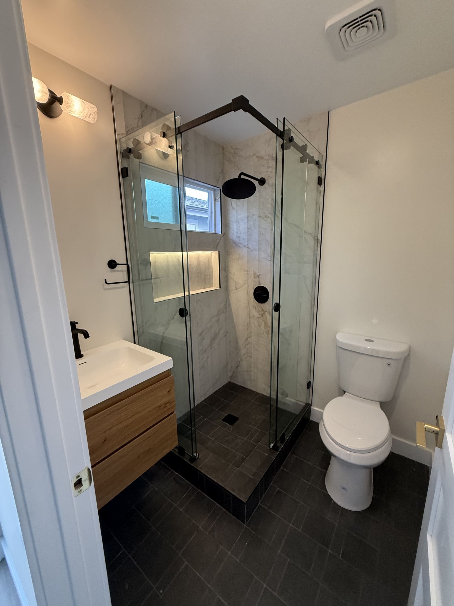 Small bathroom with glass corner shower, white toilet, sink, and dark tiled floor