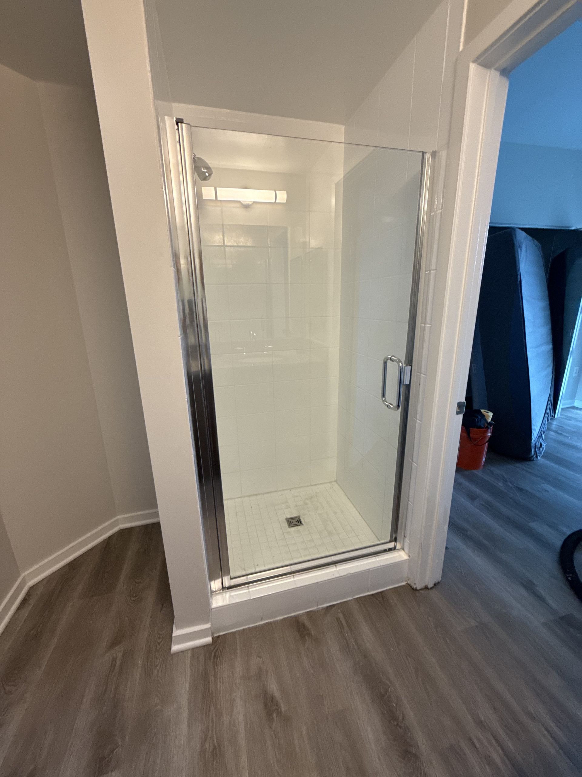 Small white-tiled shower stall with clear glass door in a bathroom with gray wood-style flooring.