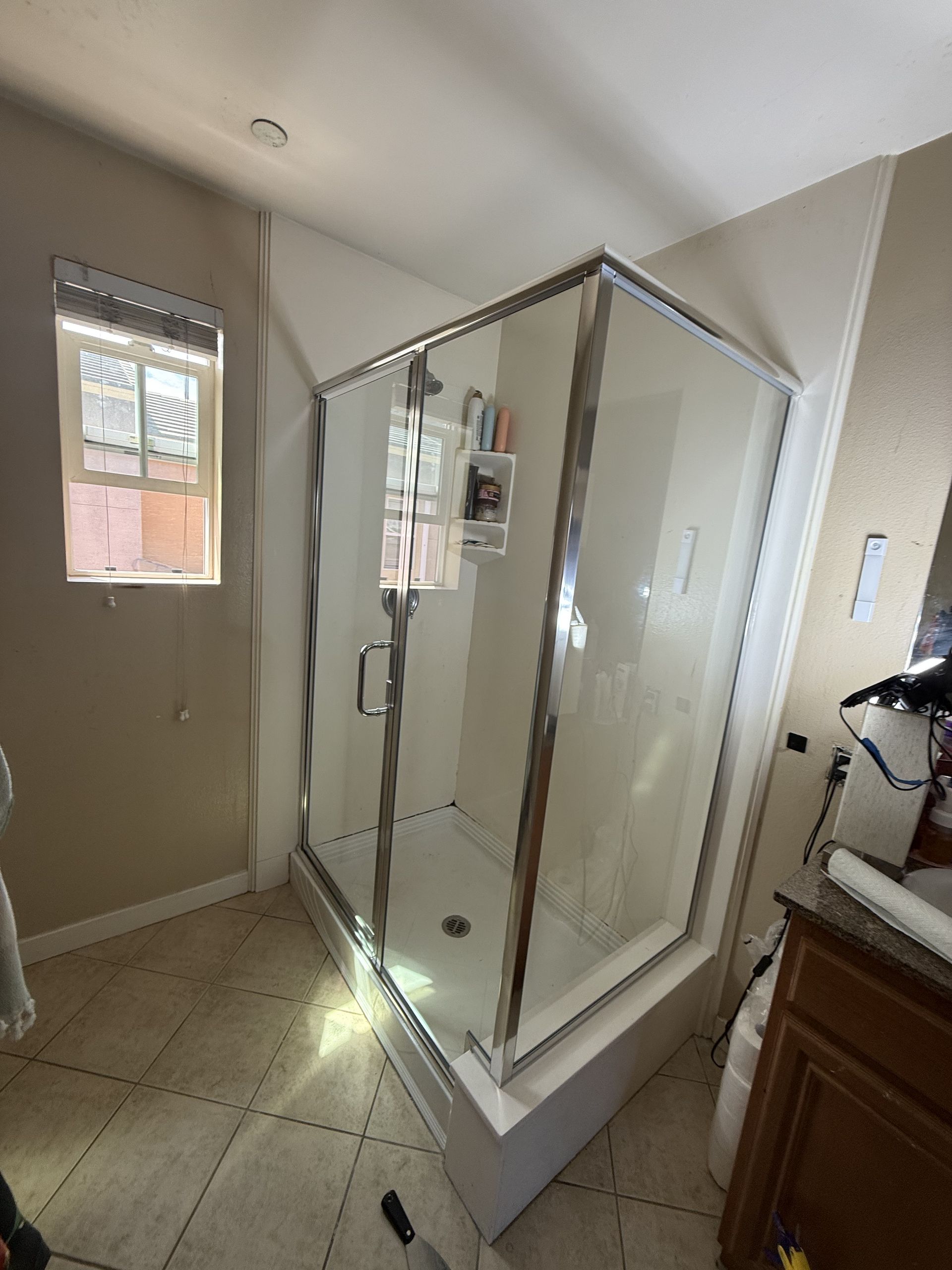 Bathroom with a glass-enclosed corner shower, beige walls, tiled floor, and a small window.