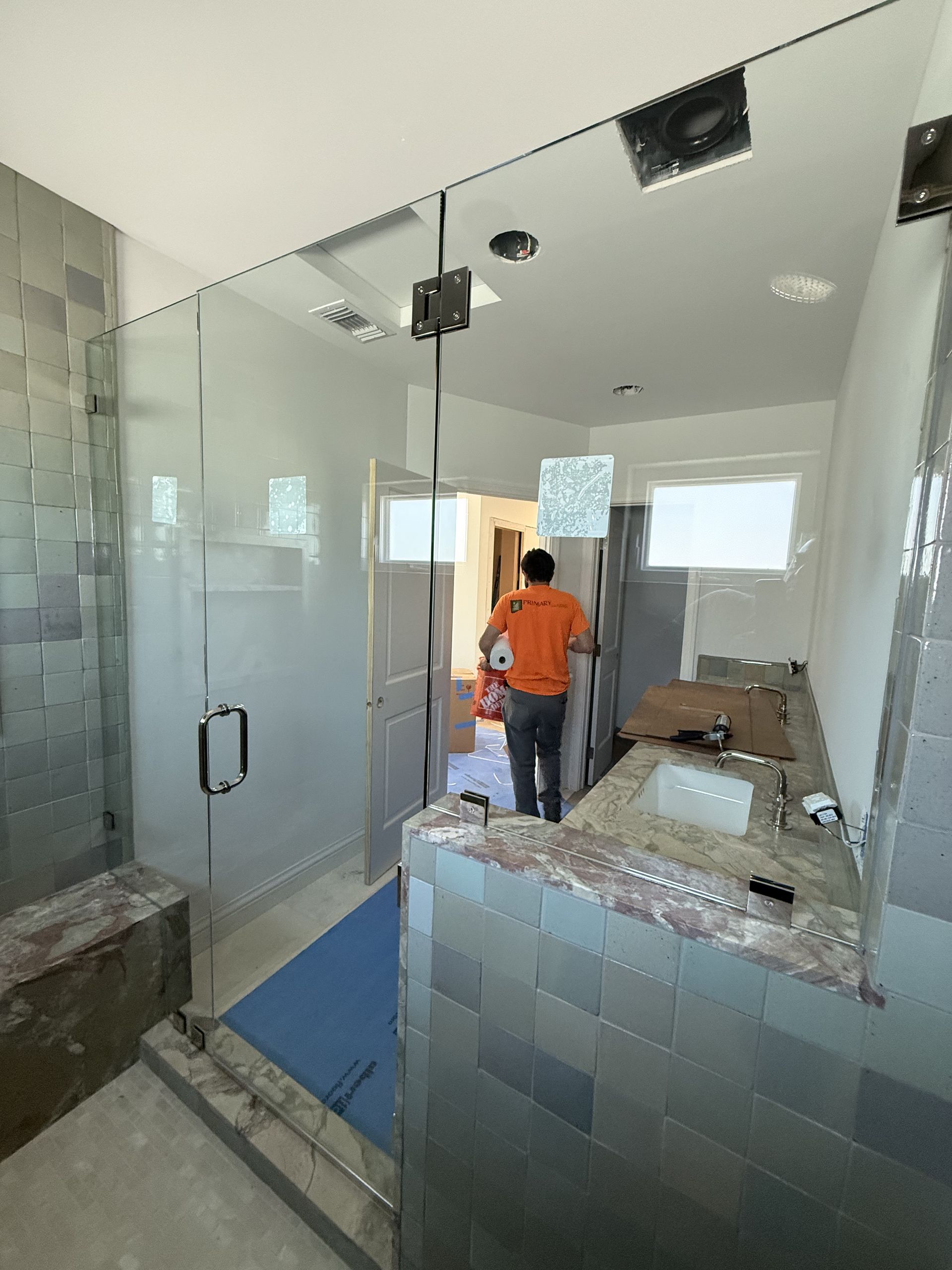 Bathroom under construction with glass shower, worker in orange shirt, and blue protective floor mat