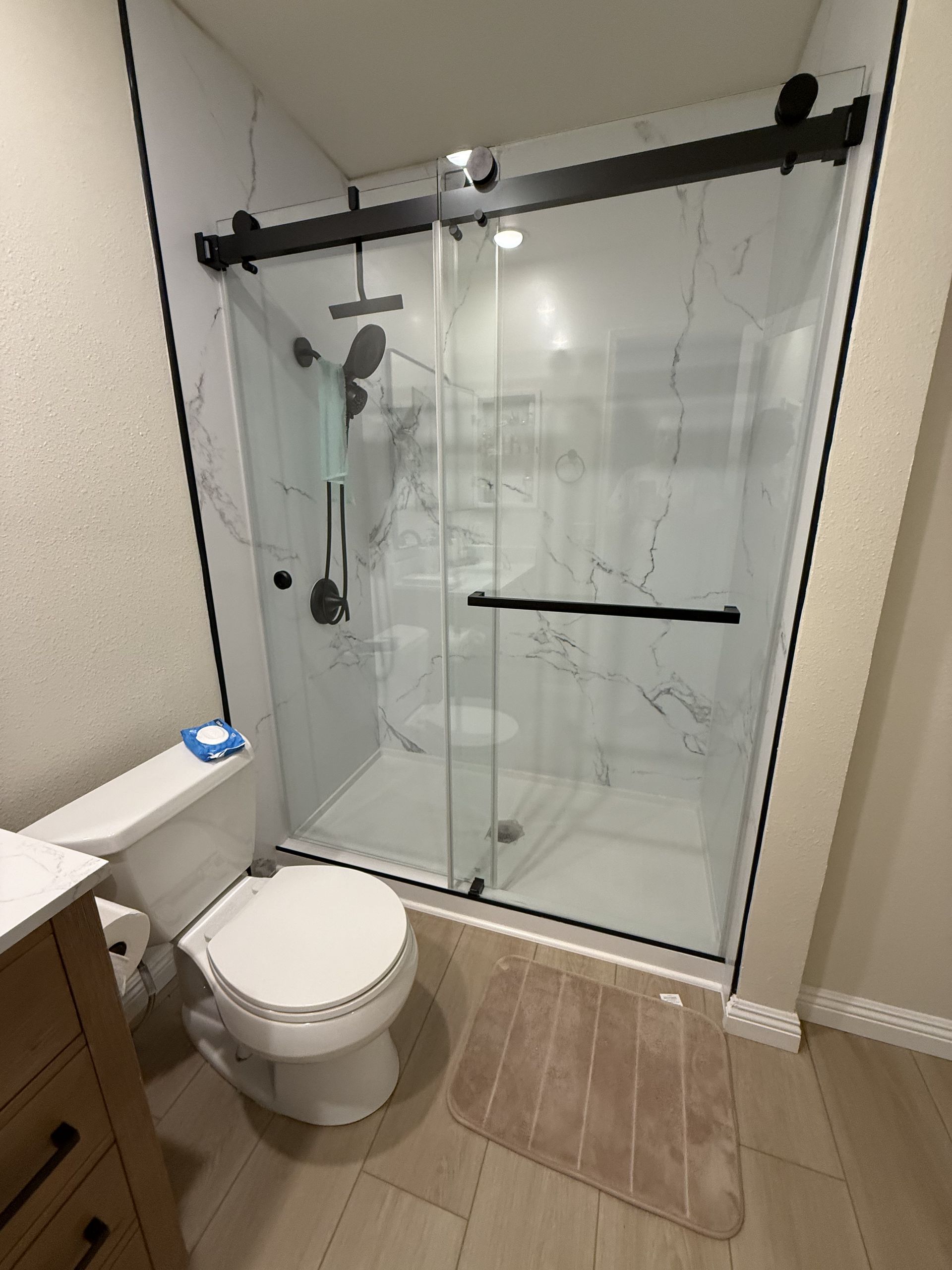 Small bathroom with toilet, beige mat, and glass shower enclosure with black hardware.
