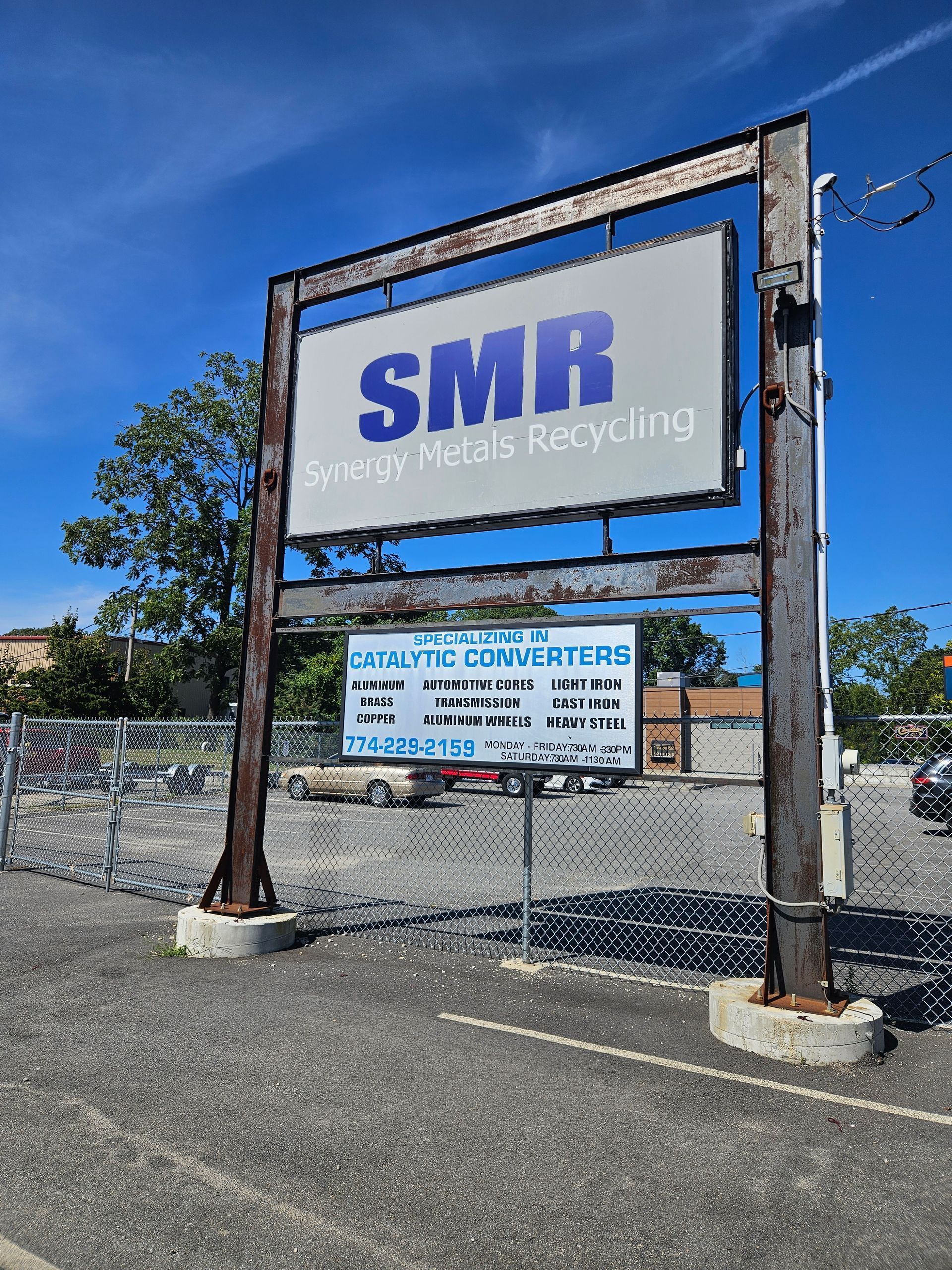 Sign for SMR Specialty Metals Recycling. Blue sky, metal signpost. Gray pavement and fence.