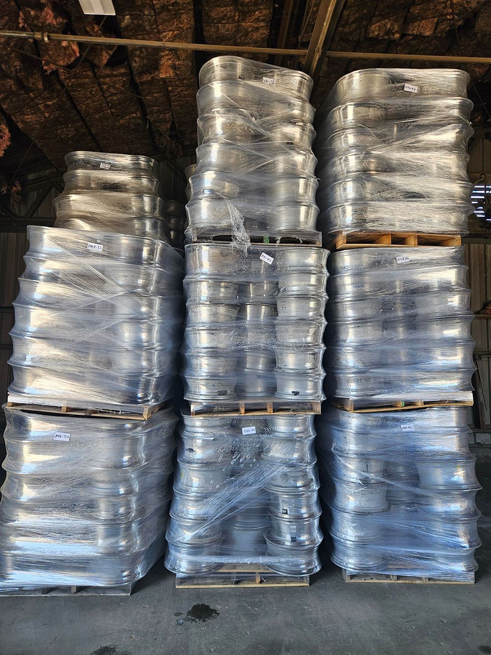 Stacks of metal kegs wrapped in plastic on wooden pallets inside a warehouse.