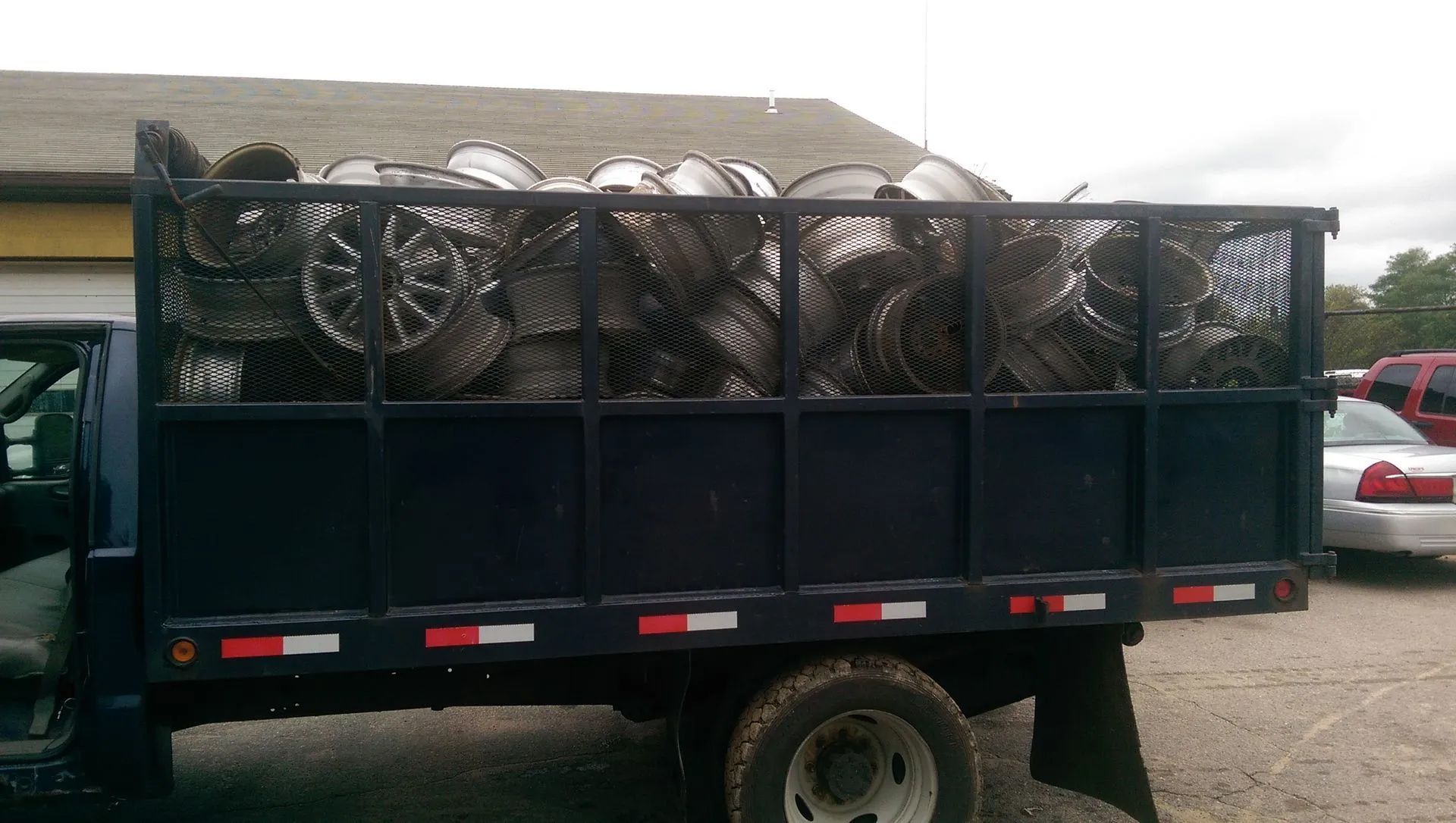 Truck bed filled with numerous silver car rims, parked outside on a cloudy day.