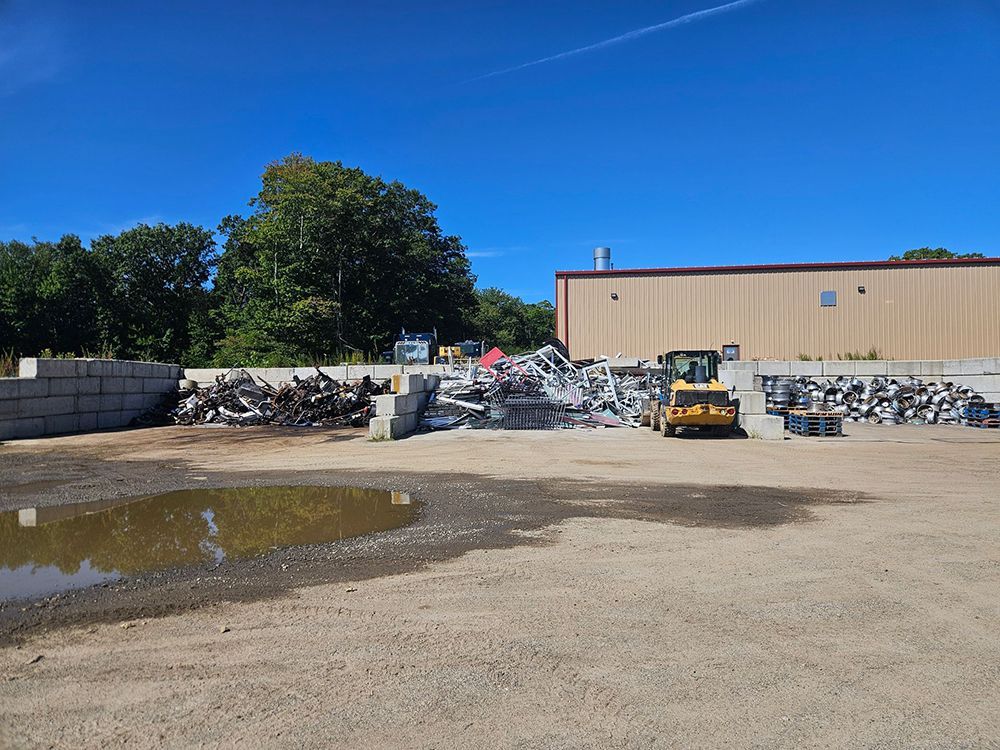 Scrap metal yard with piles of metal, a loader, and a building on a sunny day.