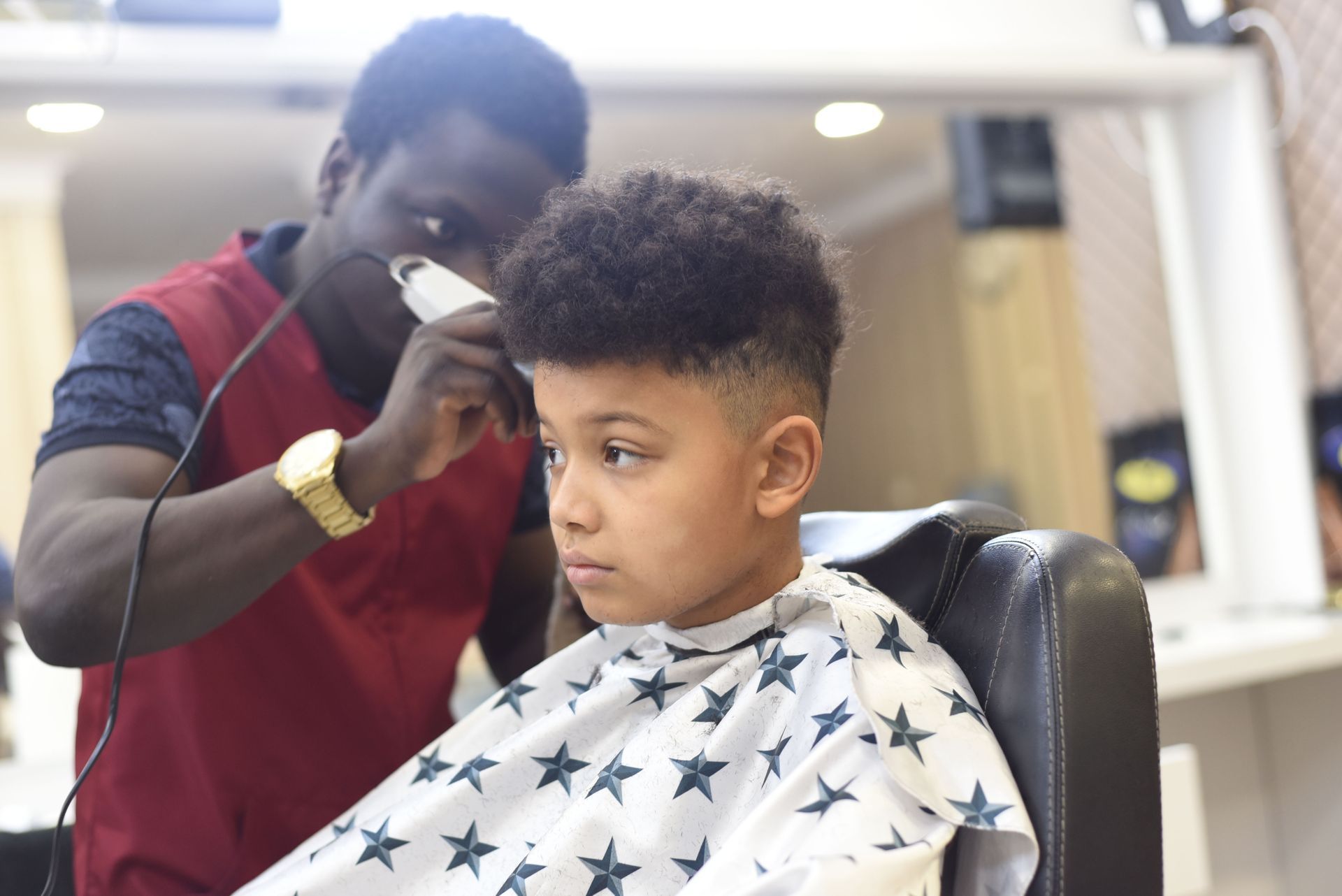 A barber in a maroon vest uses clippers to trim the hair of a person wearing a star-patterned cape in a salon.