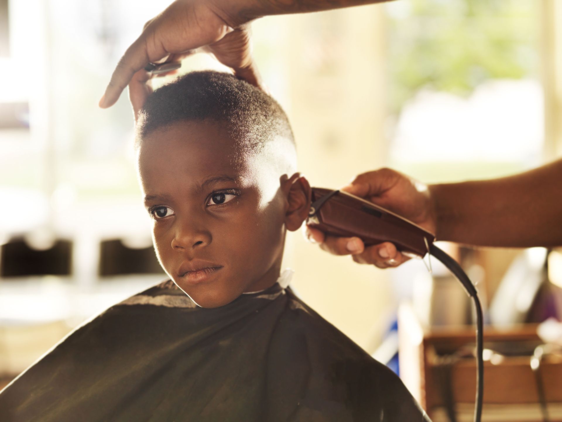 A barber uses clippers to trim the hair of a person seated in a chair, wearing a protective cape.