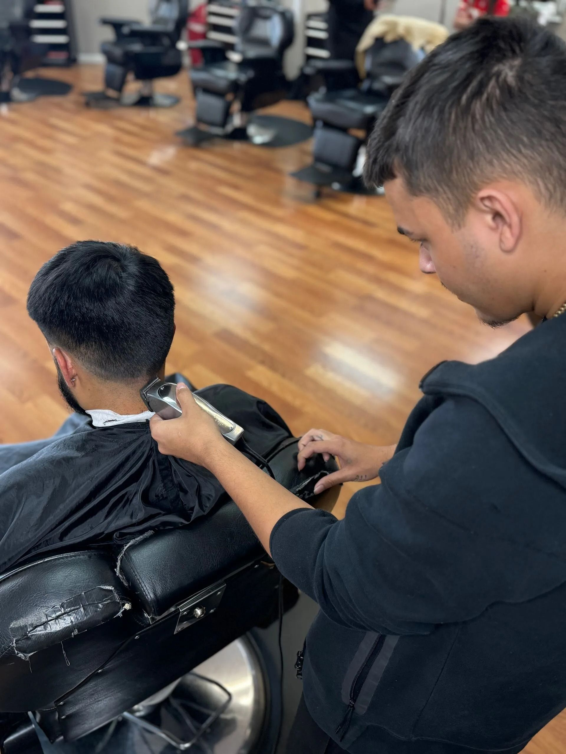 A barber in a black sweatshirt uses electric clippers to trim the neckline of a client sitting in a salon chair.