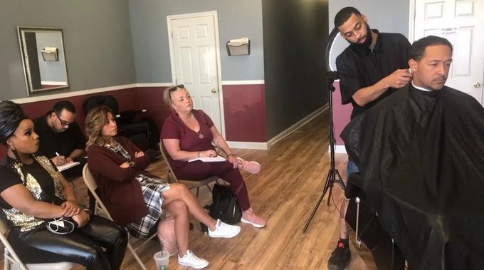 A barber gives a haircut to a client seated in a cape while three people watch from folding chairs in a shop.
