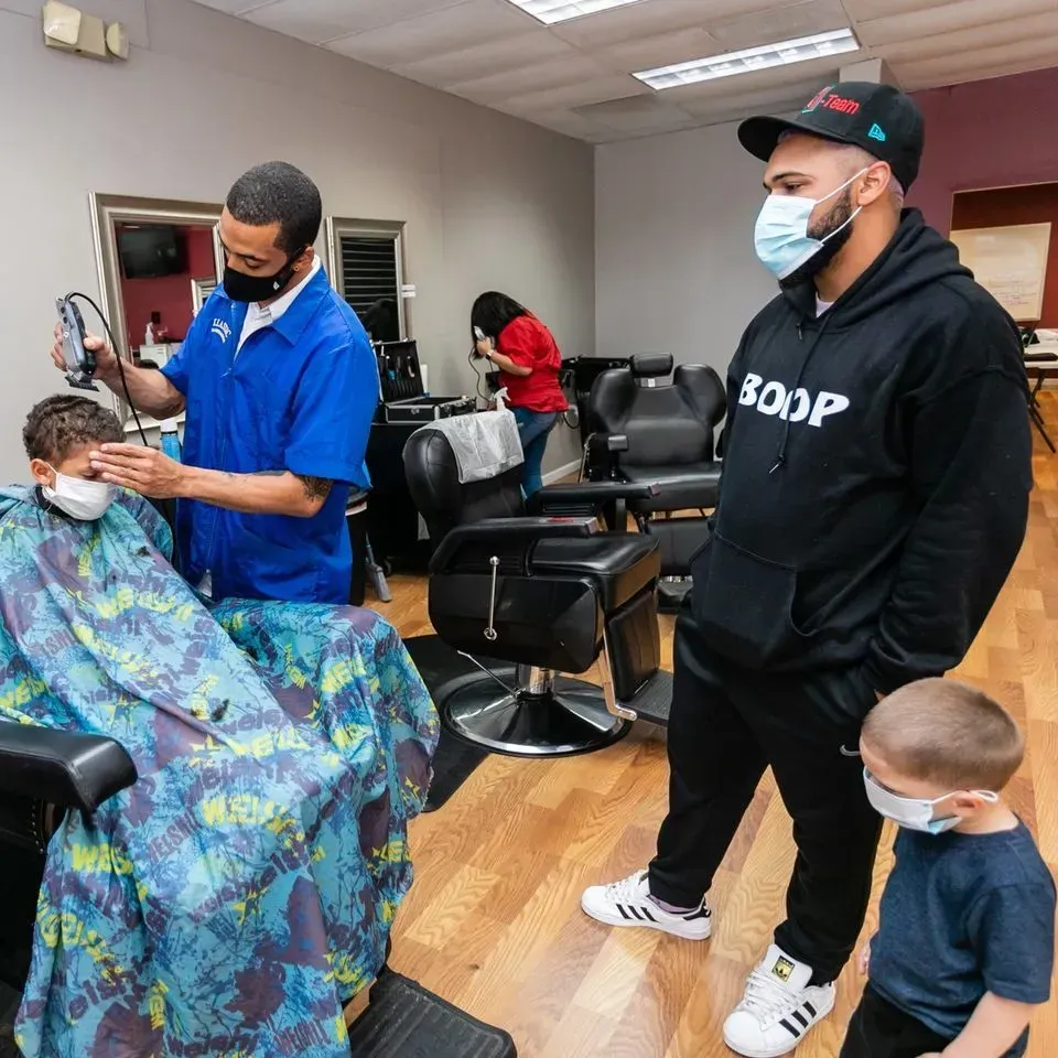A barber in a blue shirt gives a haircut to a person wearing a patterned cape, while two observers watch nearby.