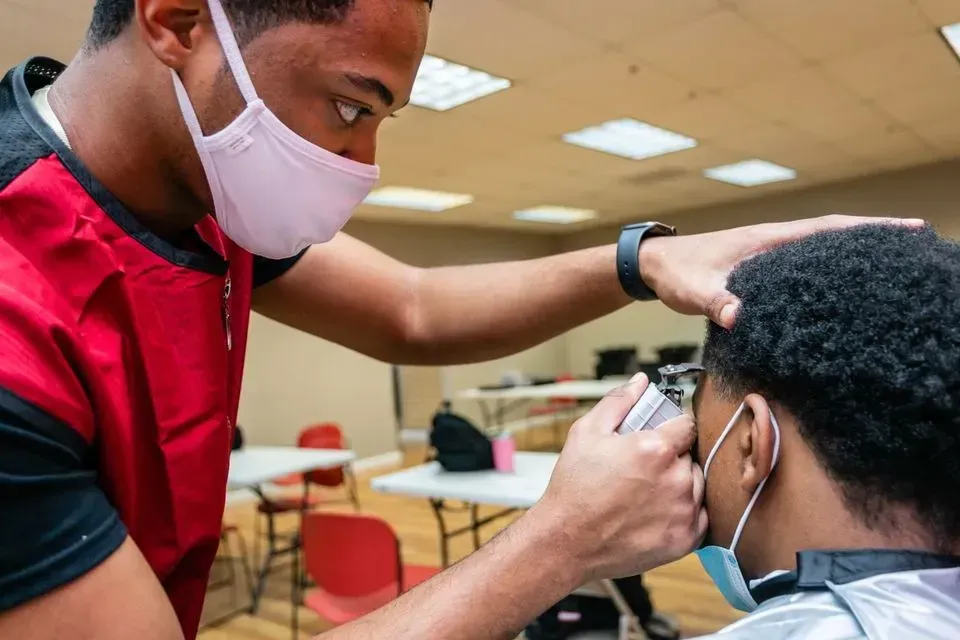 A barber wearing a red vest and pink face mask uses clippers to trim a client's hair in a room with white tables.
