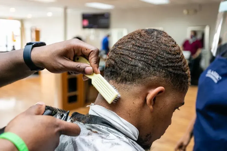 A barber uses a brush to groom a man's waves while holding electric clippers near his neckline in a barbershop.