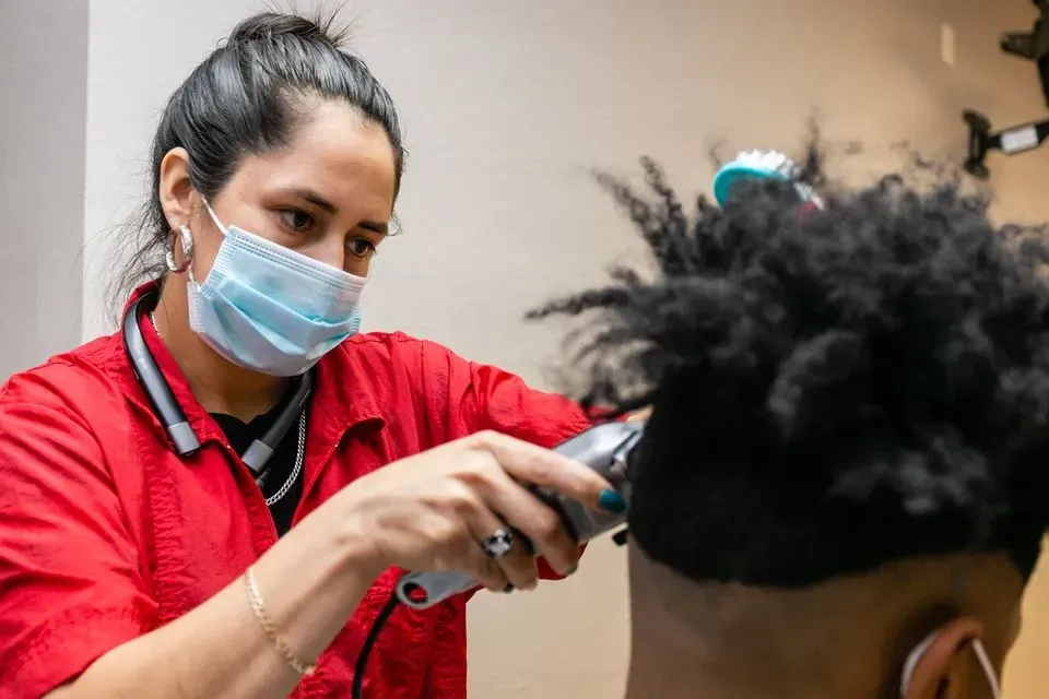 A person wearing a red shirt and face mask uses electric clippers to trim hair on the side of a client's head.