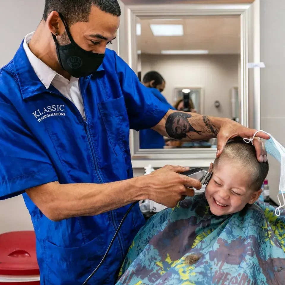 A barber in a blue uniform and face mask uses electric clippers to cut the hair of a smiling child in a colorful cape.