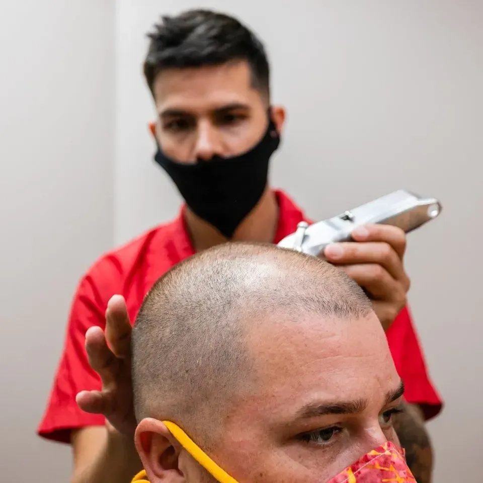 A person wearing a red shirt and black face mask uses electric clippers to cut the hair of someone wearing a mask.