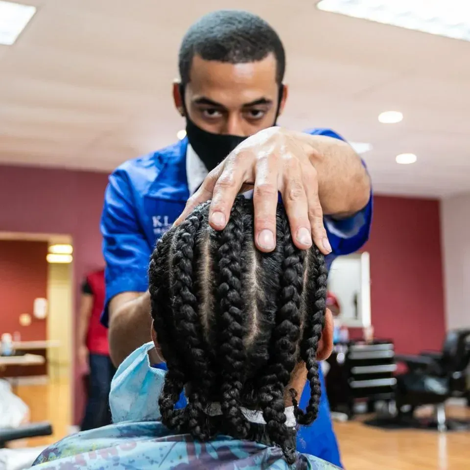 A person in a blue uniform and face mask braids another individual's hair in a salon setting.