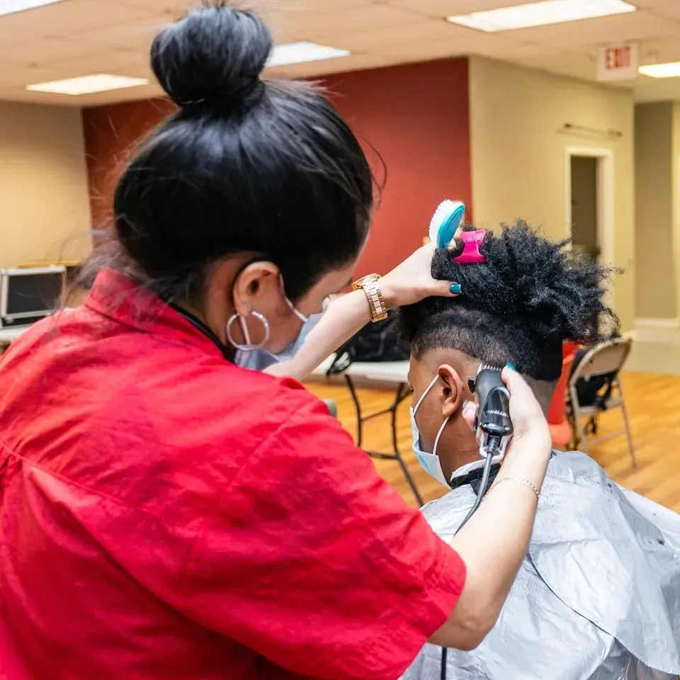 A person in a red shirt uses electric clippers to trim the hair of someone wearing a protective cape in a salon.