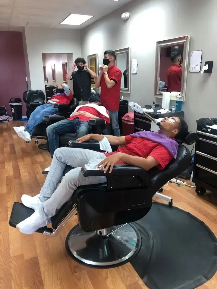 Three people in chairs in a barbershop receiving service, while two workers stand nearby.