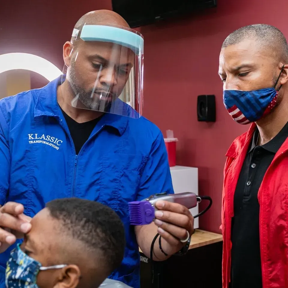 A barber wearing a face shield and blue uniform holds hair clippers while a man in a red jacket observes.