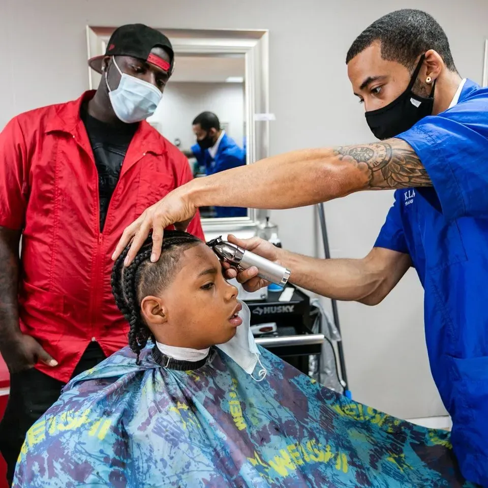A barber in a blue uniform trims a child’s hair while a person in a red shirt watches in a brightly lit shop.