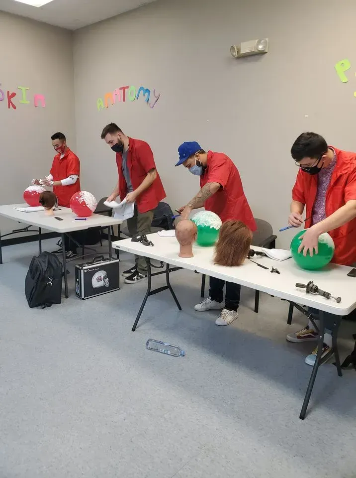 Four individuals in matching red jackets work at tables with mannequin heads and craft materials in a classroom setting.