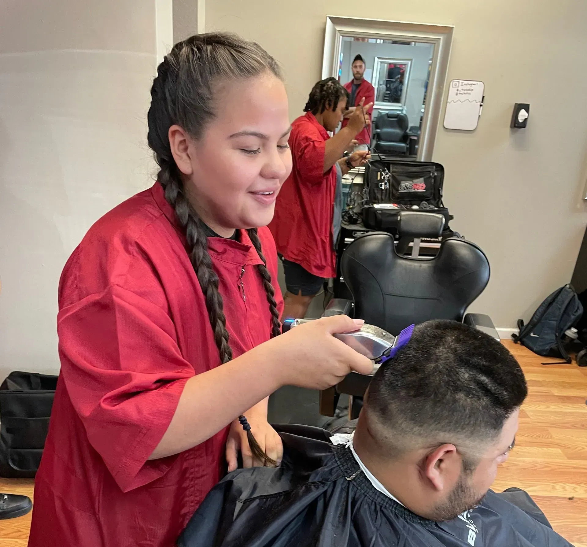 A stylist in a red smock uses clippers to trim the hair of a client in a barber chair inside a salon.