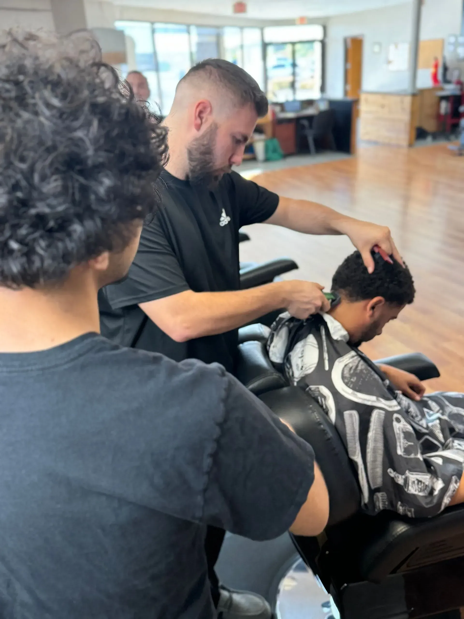 A barber in a black t-shirt uses clippers to trim a client's hair in a brightly lit shop.