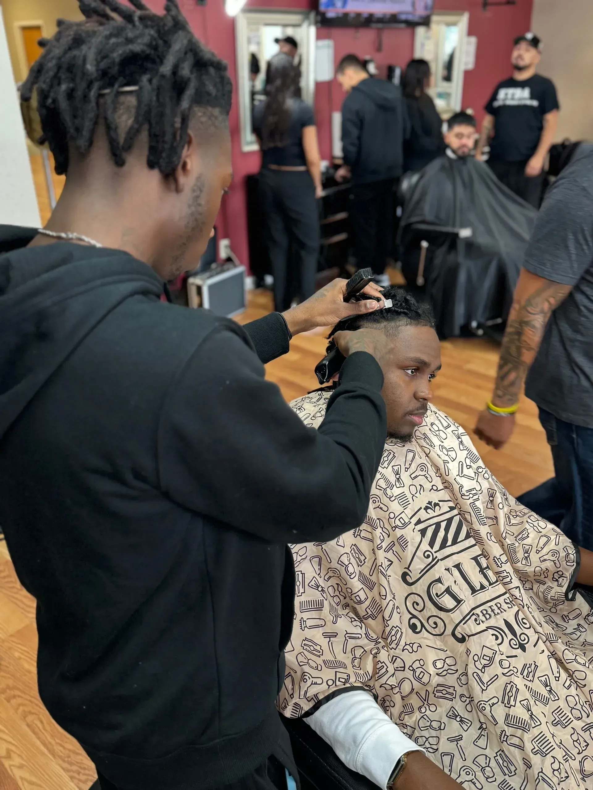A barber in a black hoodie grooms a customer's hair in a brightly lit barbershop.