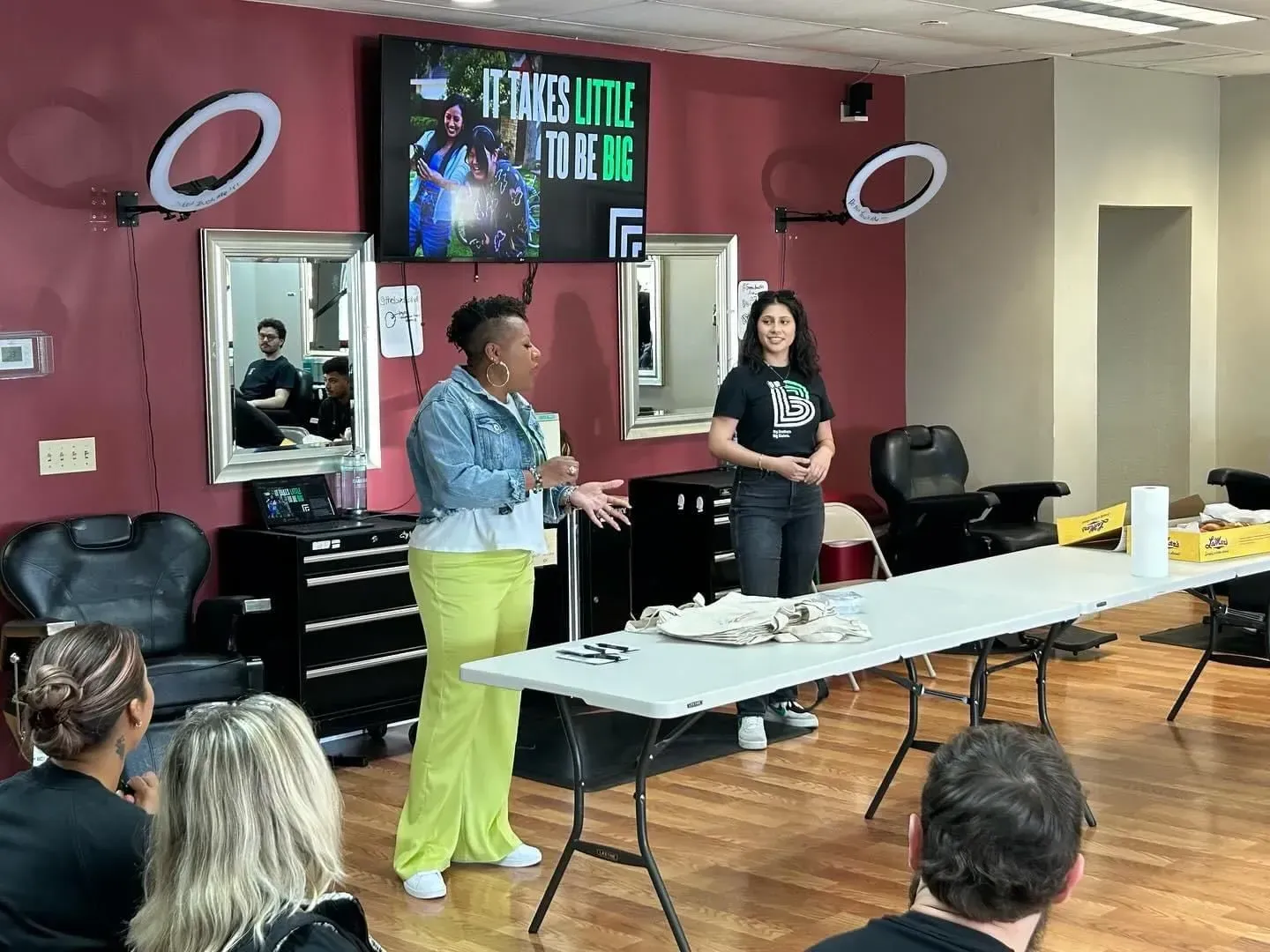 A presenter in a denim jacket and yellow pants speaks to a seated audience in a salon, with another person standing nearby.