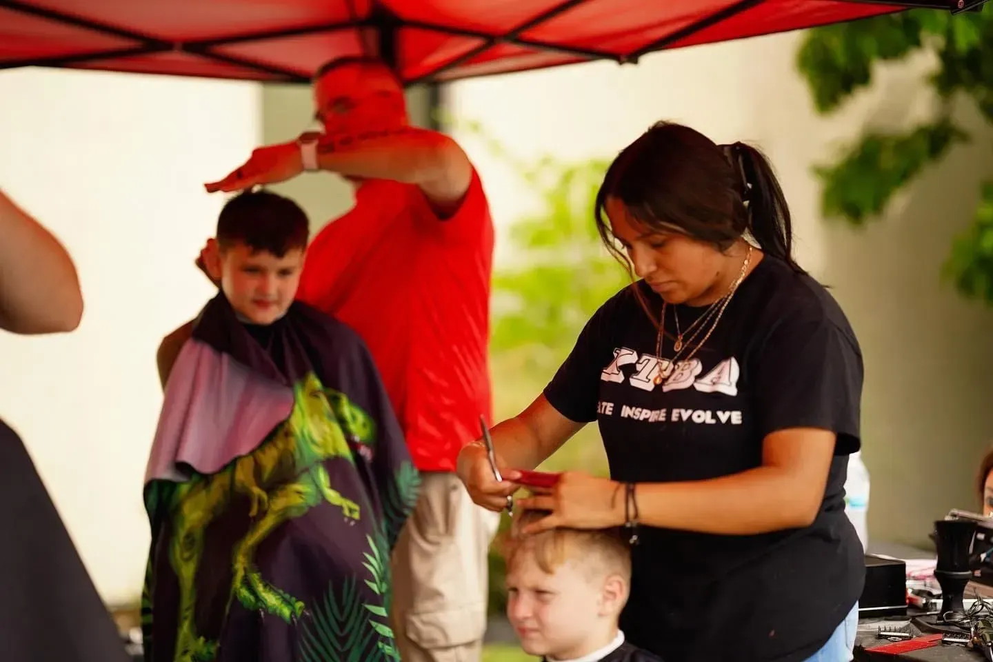 Two barbers provide haircuts to children under a red canopy tent.