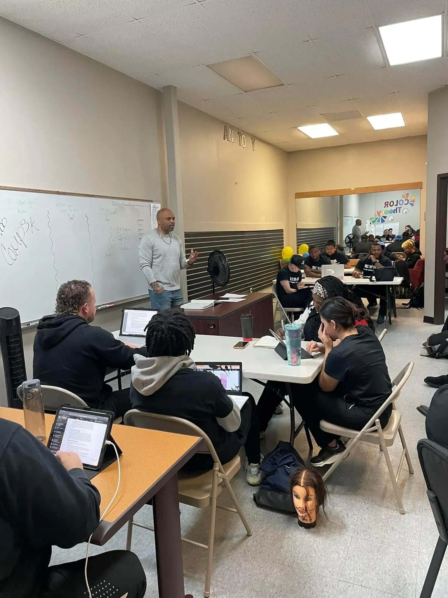 A classroom with students at tables and a teacher standing near a whiteboard, facilitating a group lesson.