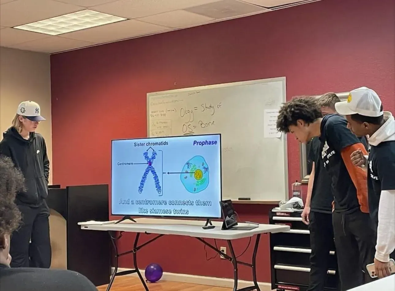 A group of students stands in a classroom watching a digital presentation about cell structure on a television monitor.