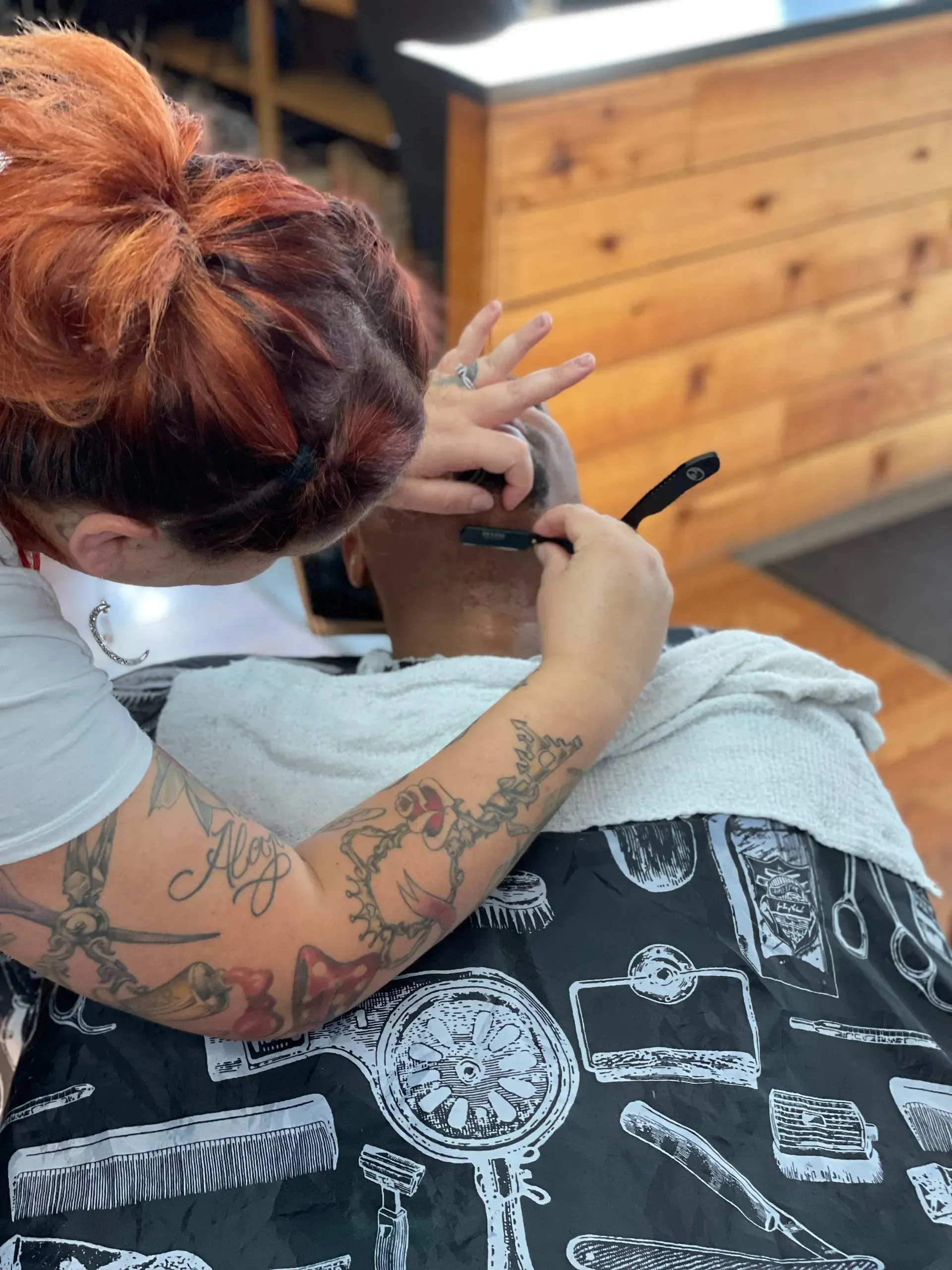 A barber with tattooed arms uses a straight razor to shave the back of a client's neck in a shop.