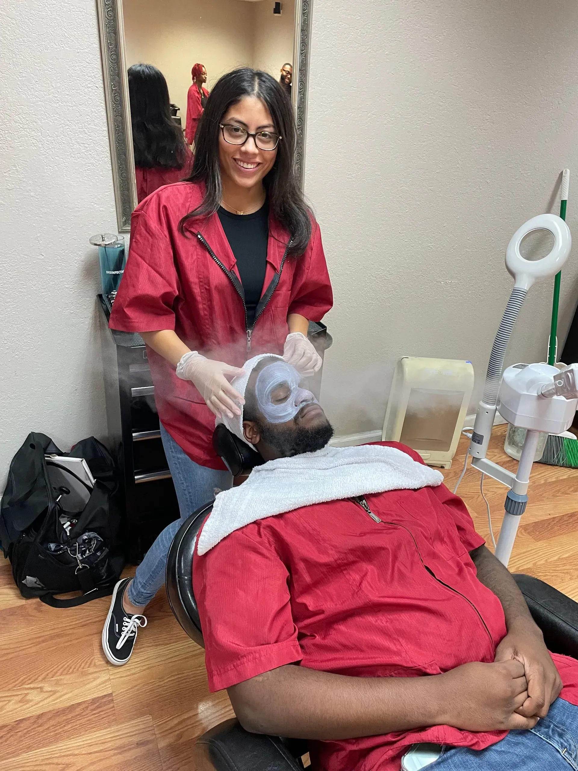 An esthetician in a red smock performs a facial with steam on a client sitting in a salon chair.