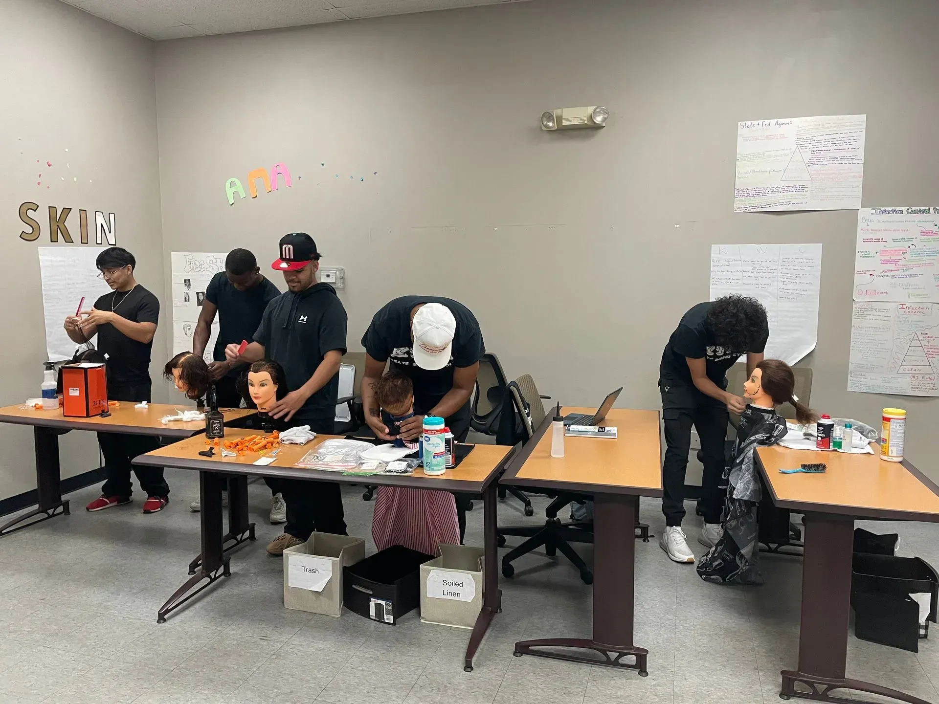 Students in a classroom practice haircutting techniques on mannequin heads at individual tables.