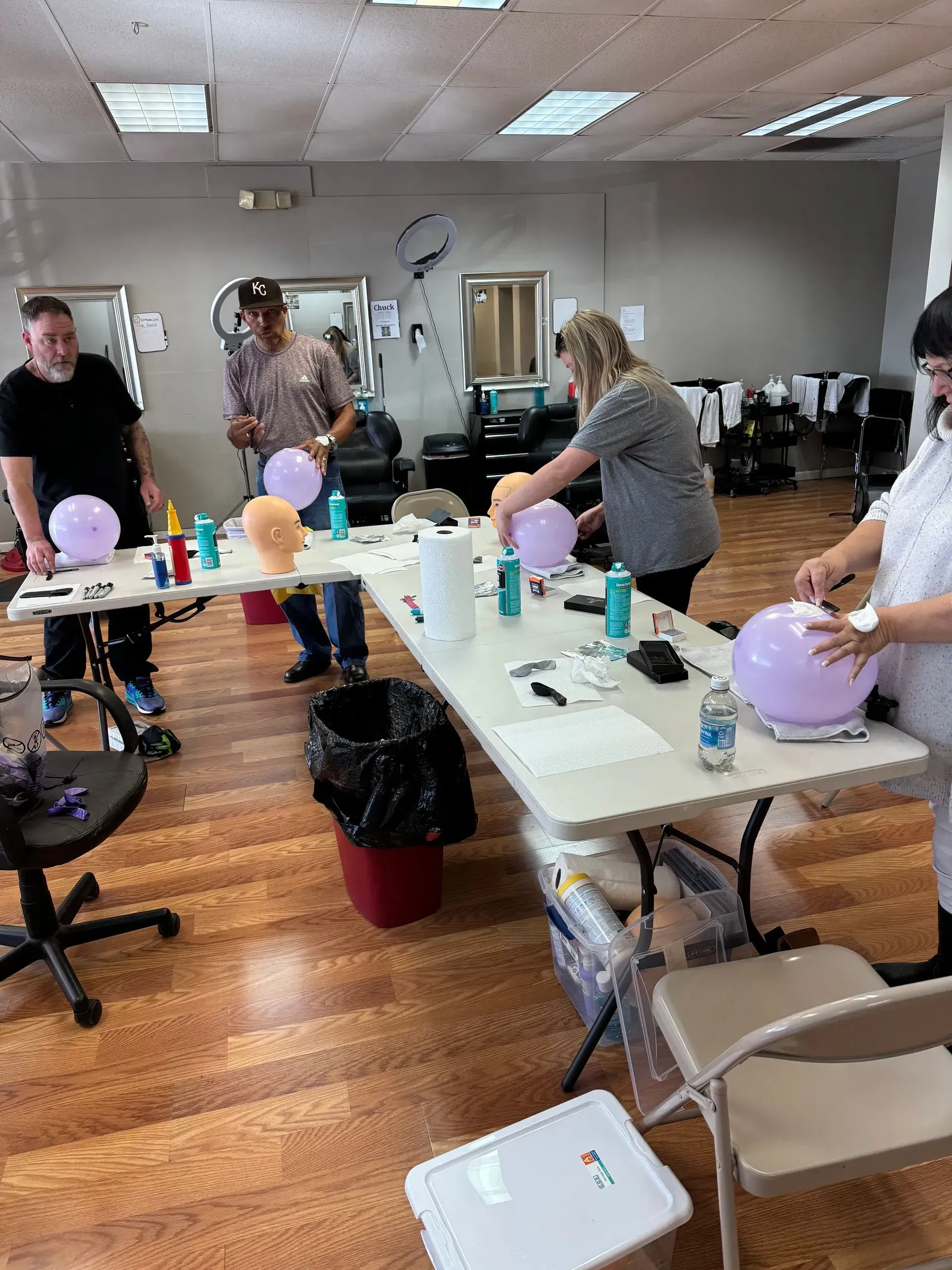 Four people stand around a table in a salon, working with lavender balloons and crafting supplies.