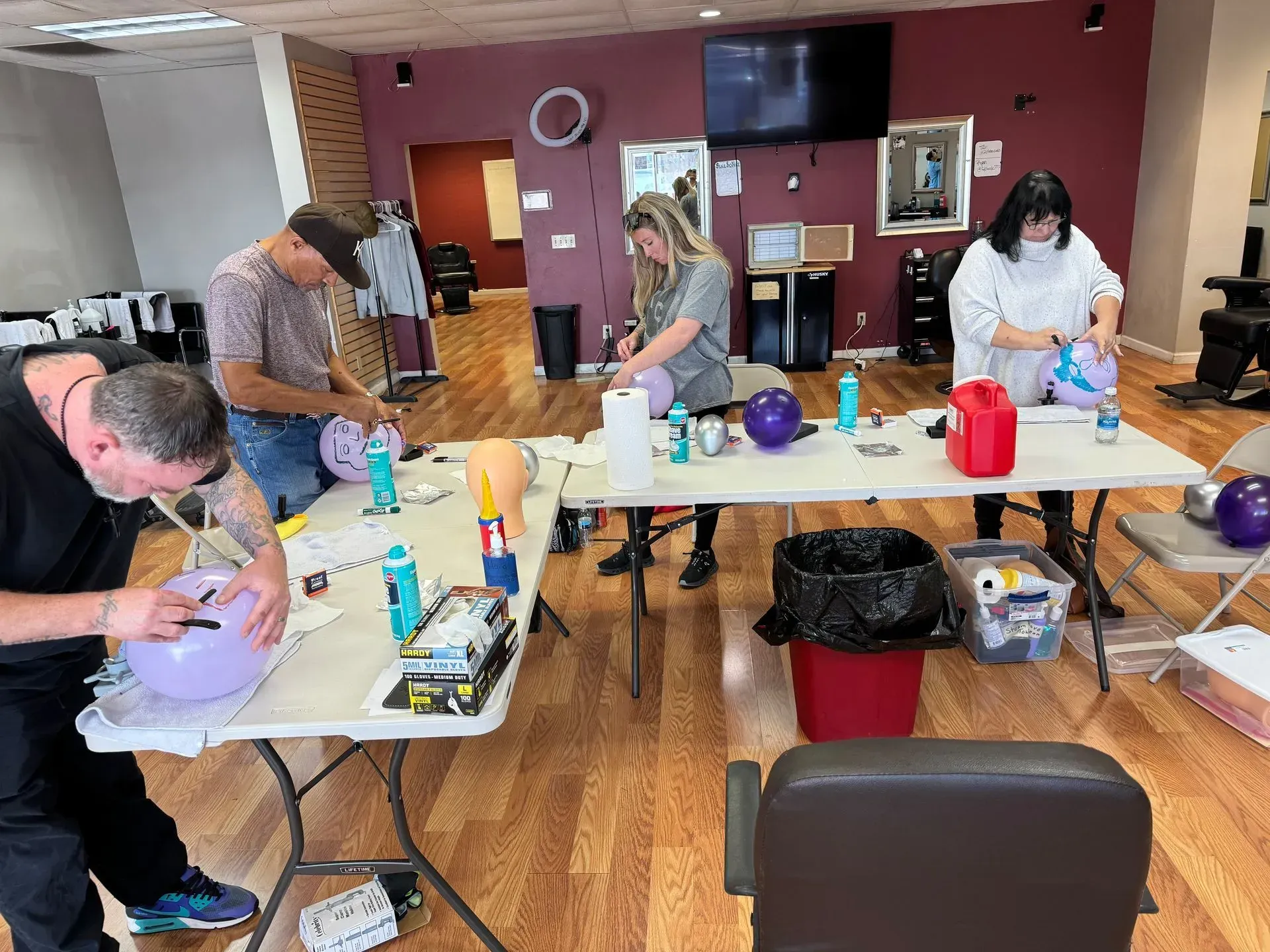 Three people work at tables in a room, decorating balloons with markers and supplies to prepare for an event.