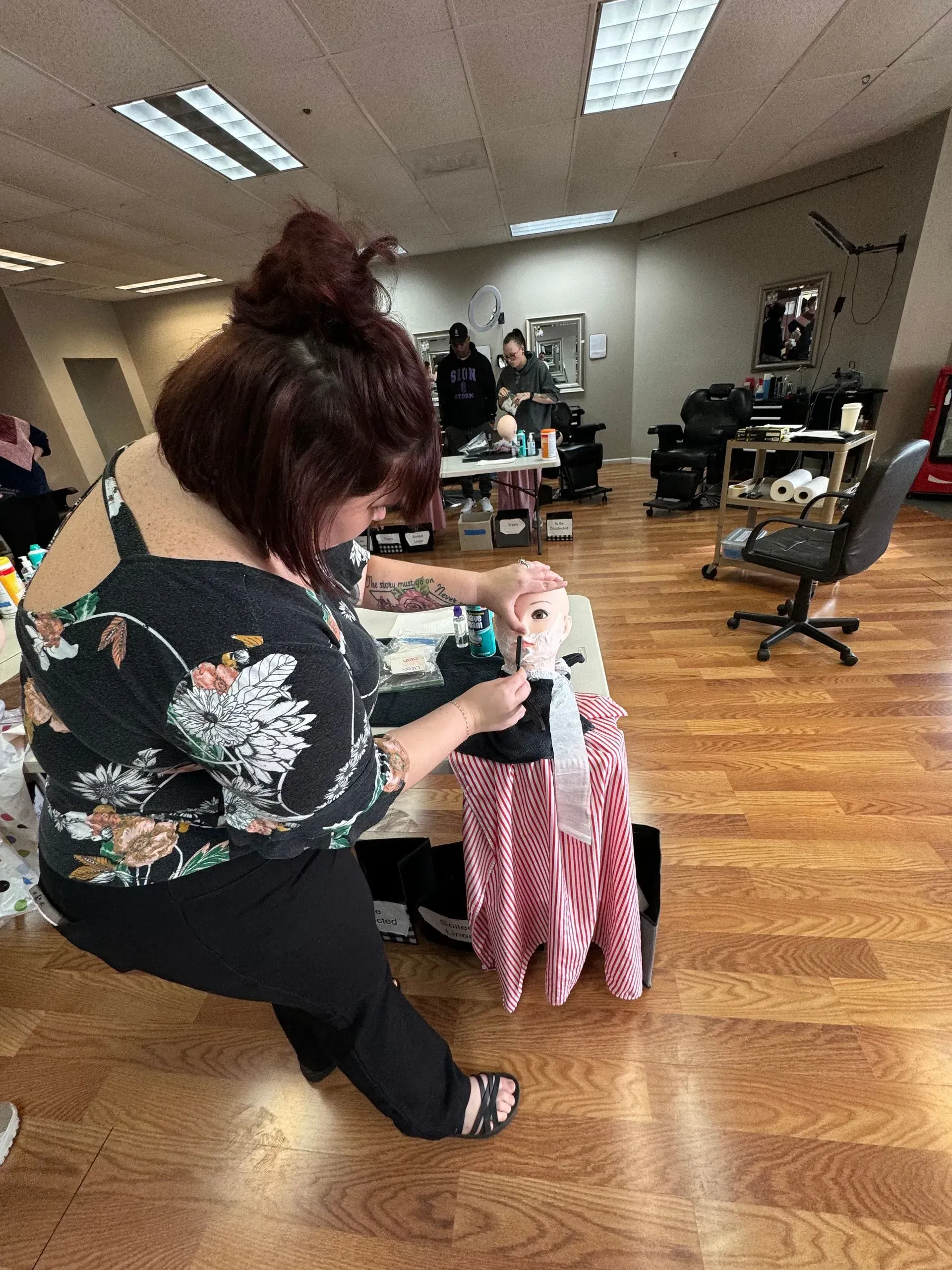 A person with auburn hair in a floral top cuts the hair of a mannequin head on a table in a bright, open salon.
