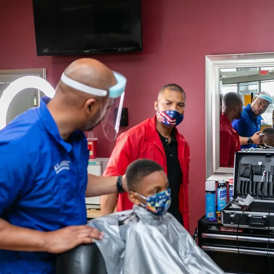 A barber wearing a face shield cuts a young person's hair in a salon while a person in a red jacket watches.