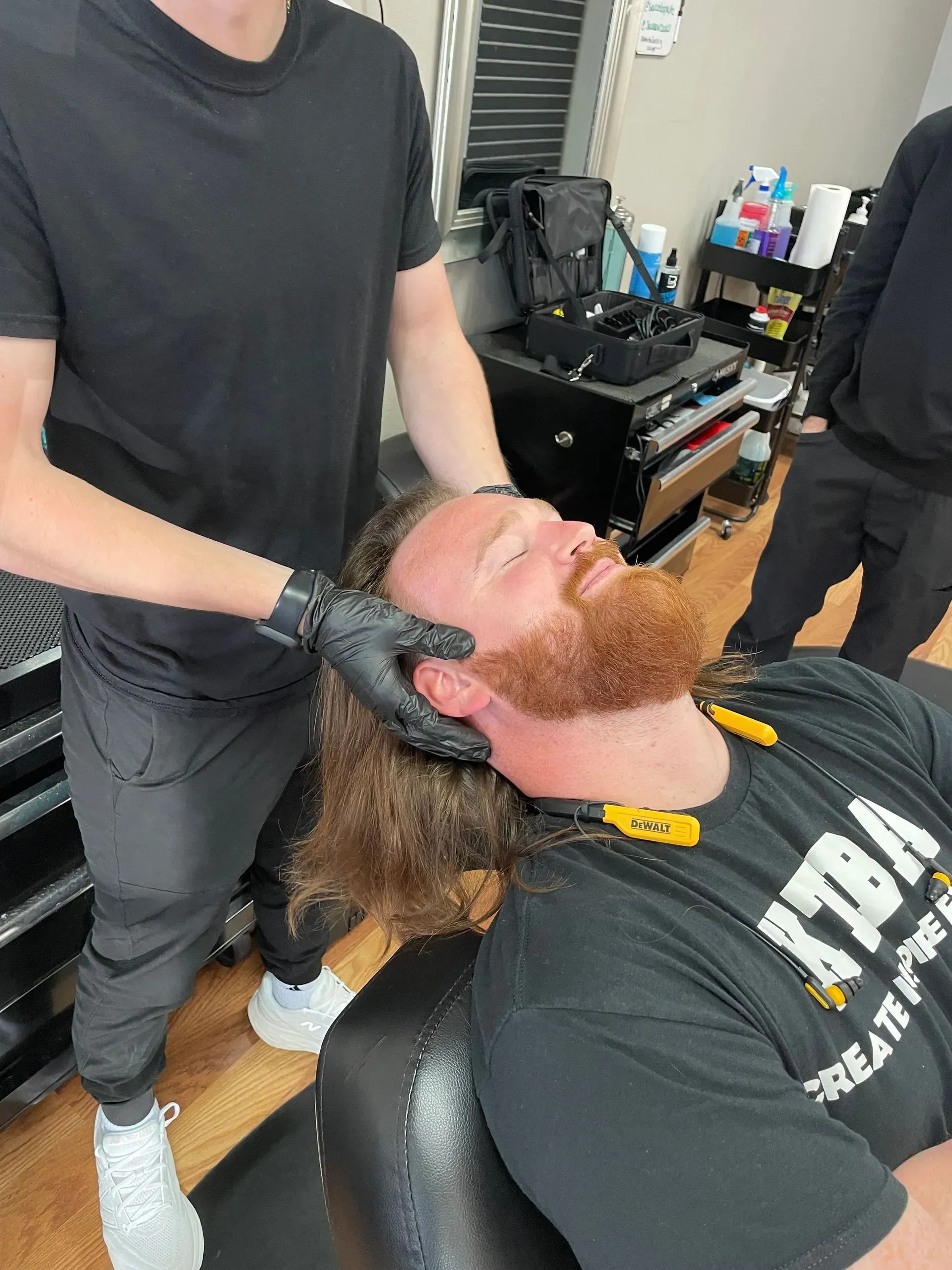 A person wearing black gloves adjusts the head of a seated individual in a salon or barbershop setting.