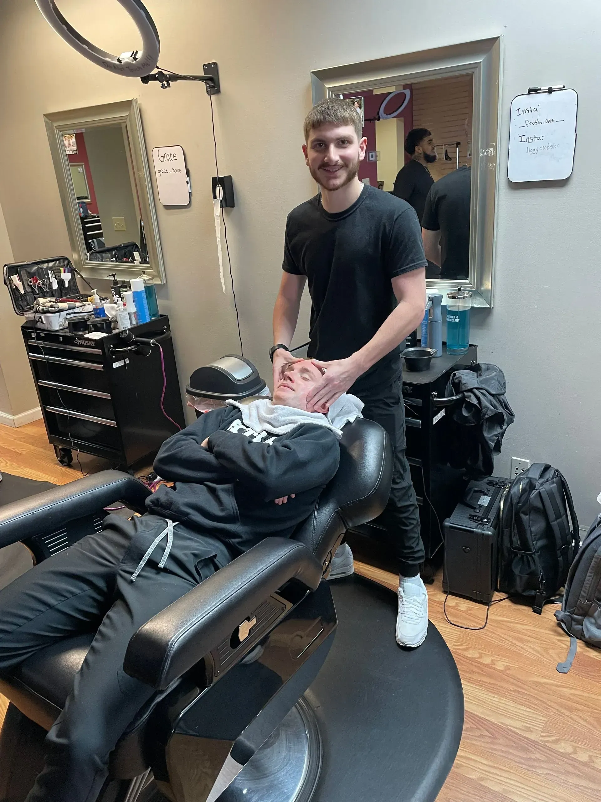 A barber stands behind a customer in a chair, applying a treatment to the person's face inside a salon.