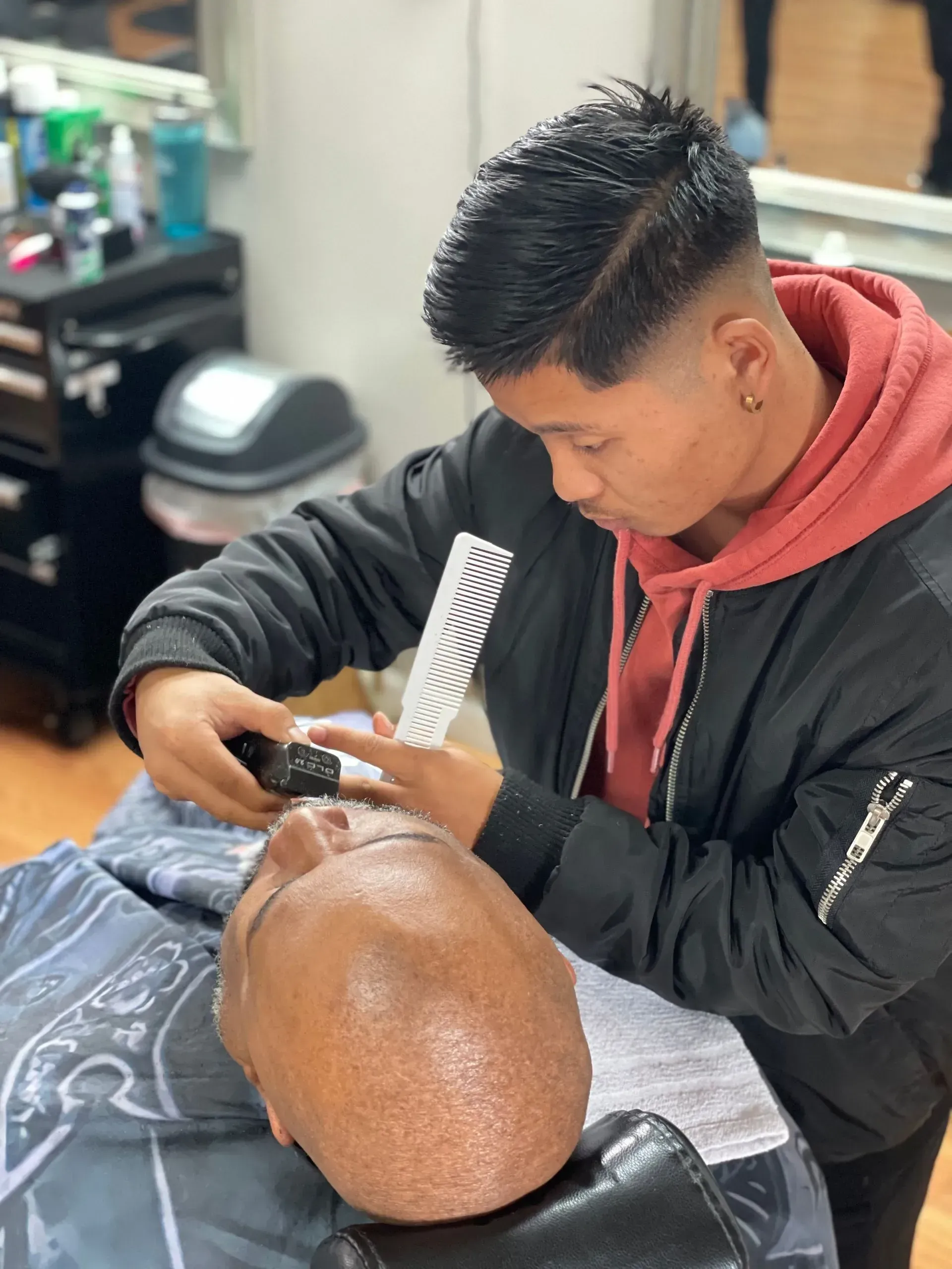 A person in a black jacket and red hoodie trims a customer's hair with clippers and a comb in a barbershop.