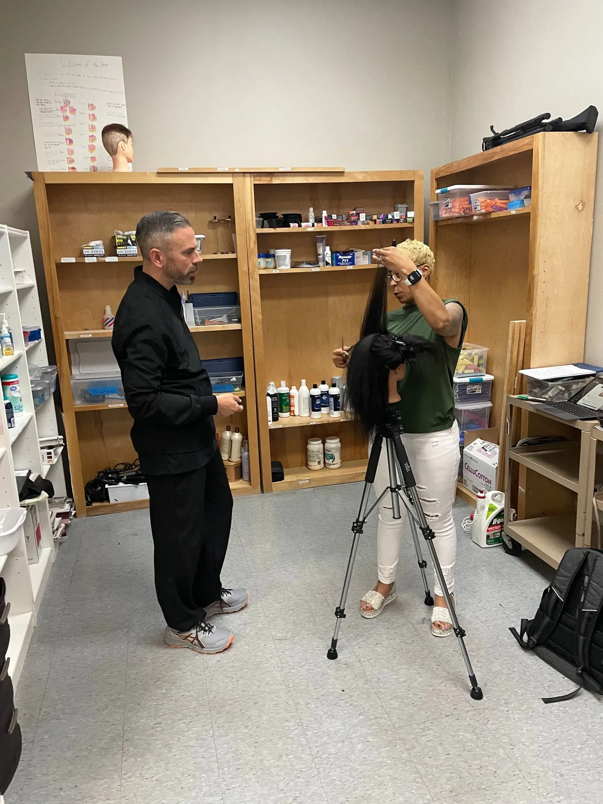 An instructor observes a student practicing hair styling on a mannequin head mounted on a tripod in a salon classroom.