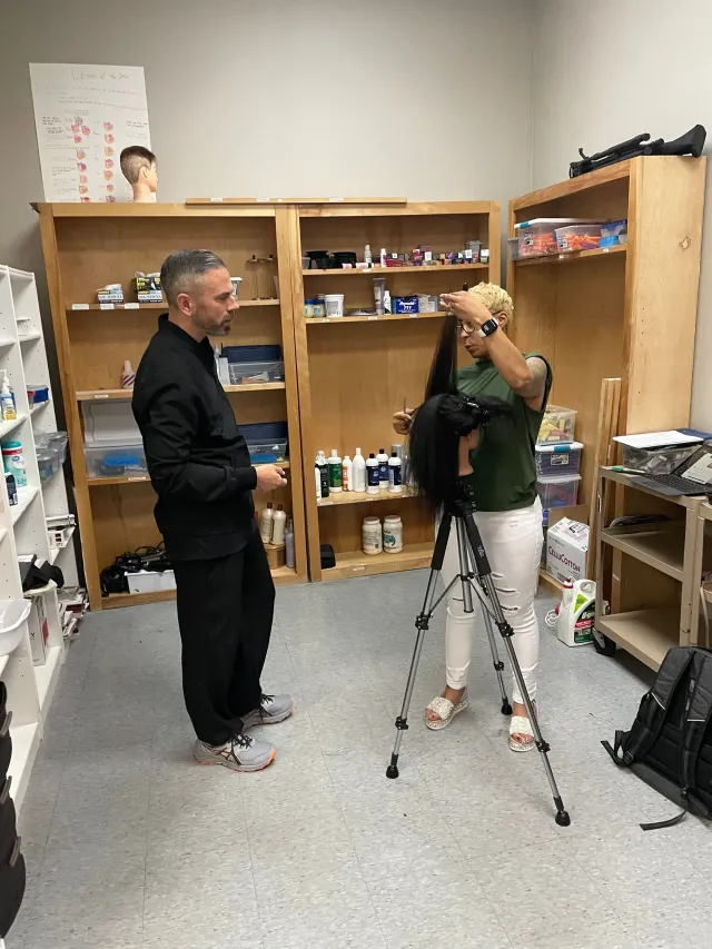 A person instructing another on styling a mannequin head mounted on a tripod inside a classroom with supply shelves.