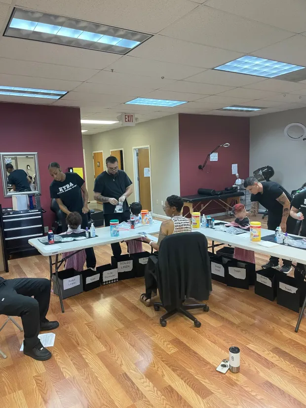 Barbers in a workshop setting work on clients seated at folding tables, with equipment and chairs throughout the room.