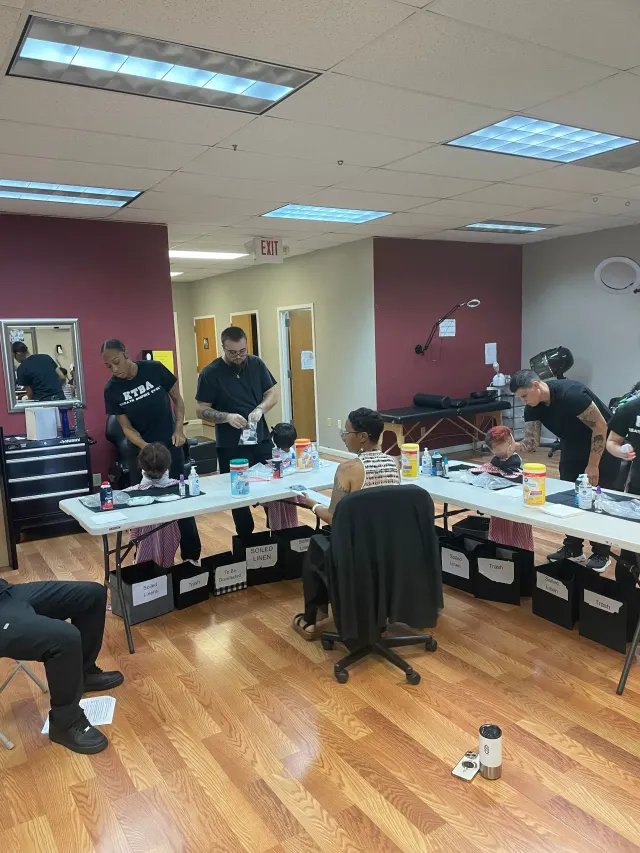 Barbers in a workshop setting work on clients seated at folding tables, with equipment and chairs throughout the room.