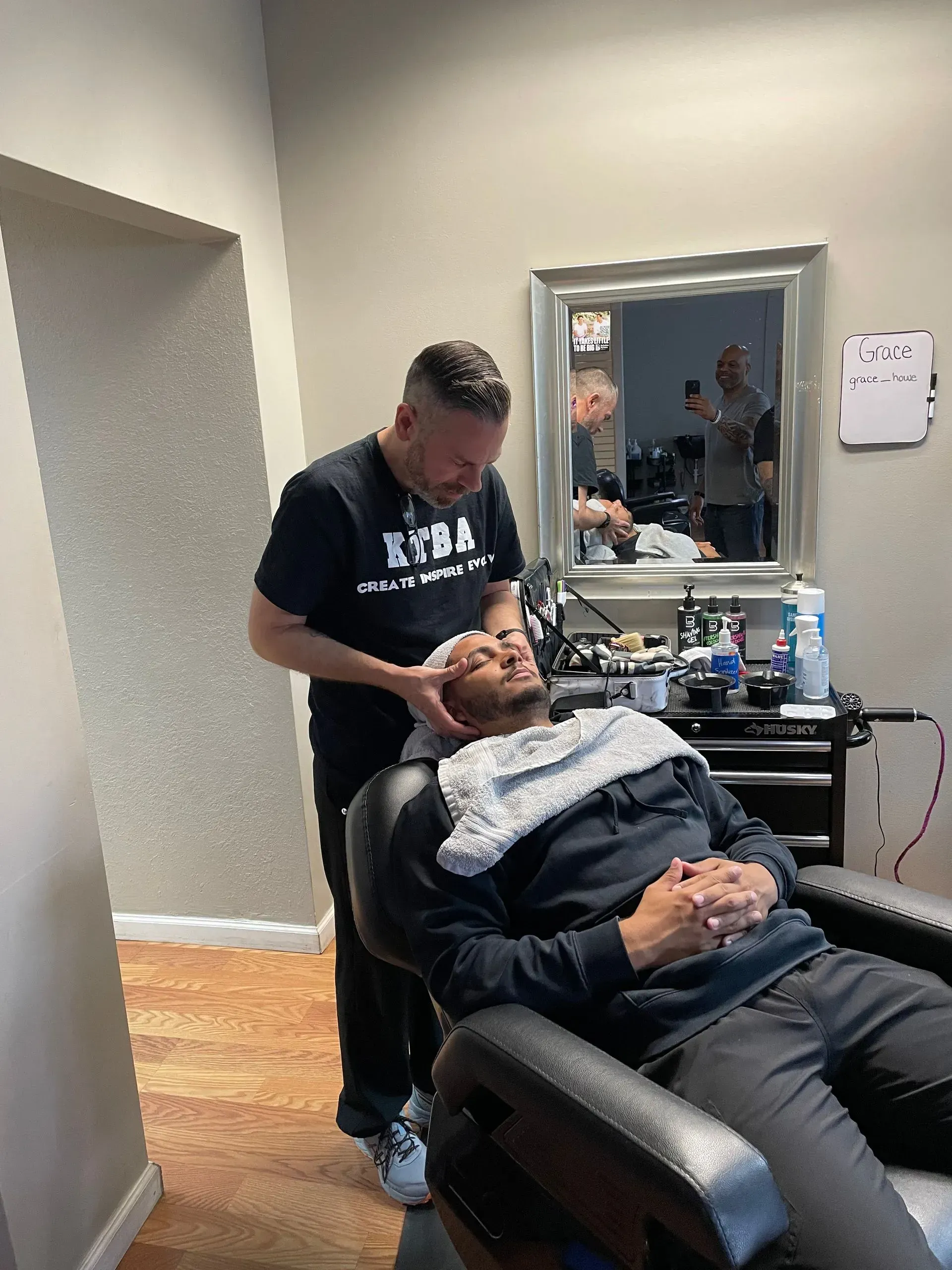 A barber provides a service to a client reclined in a salon chair, with a towel draped over the client's shoulders.